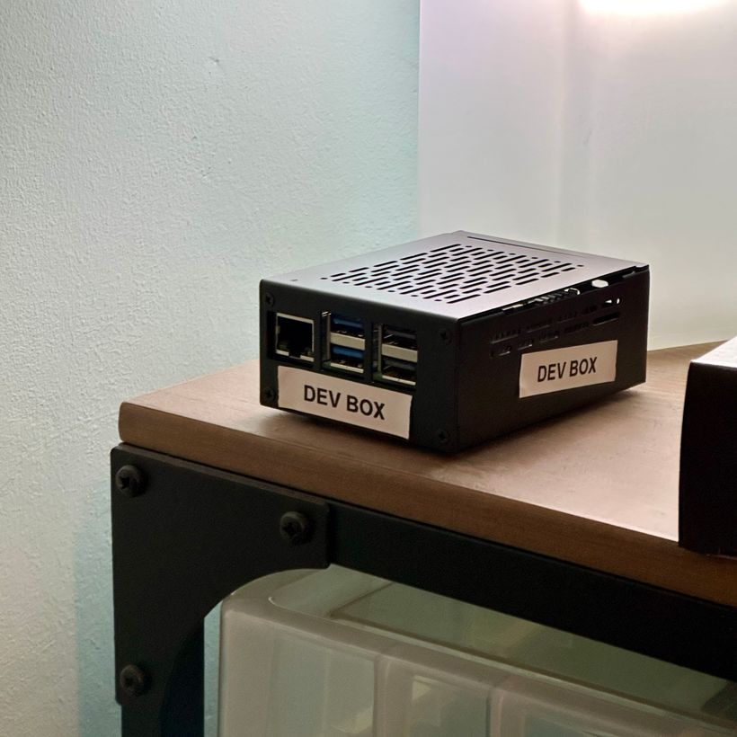 A small black metal computer case labeled 'DEV BOX' sits on a wooden surface. The case has ventilation holes on top and visible USB and ethernet ports on the front, indicating it is a Raspberry Pi inside the case.