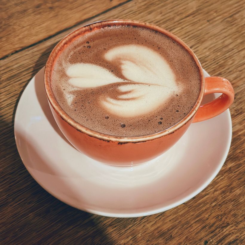 A ceramic cup filled with frothy coffee sits on a white saucer. The surface of the coffee features intricately crafted latte art resembling two leaves. The cup has a soft orange hue and is placed on a wooden table, enhancing the cozy atmosphere.