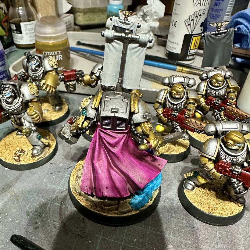 A group of space marine miniatures arranged around a central figure, which is viewed from the back, to show off a flowing robe painted a deep magenta colour. The figures are all wearing heavy silver armour with gold accents, and holding various weapons. The workspace is cluttered with paints and tools, suggesting an active model creation or painting process.