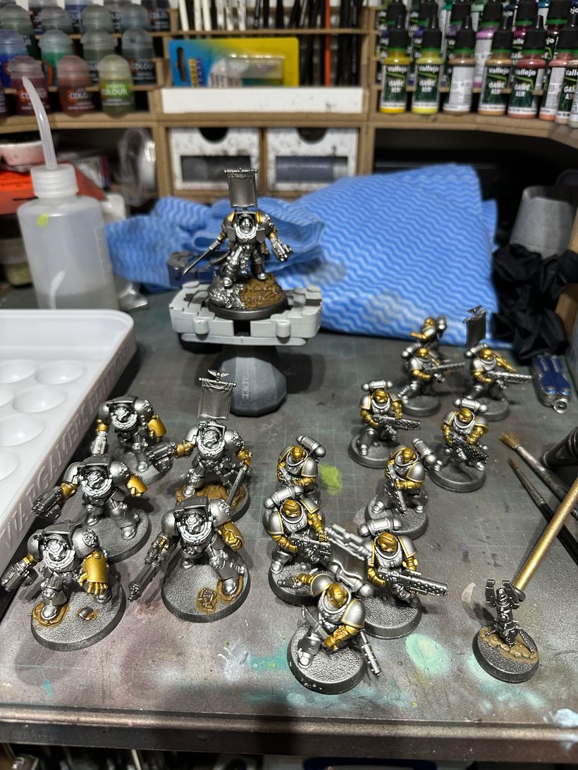 A miniatures painting workspace features a prominent silver and gold Space Marine on a raised base, holding weapons. Surrounding it are numerous smaller Space Marine figures, primarily silver, with some gold accents. The background displays paint bottles and tools, with a blue cloth and a palette visible on the workbench.