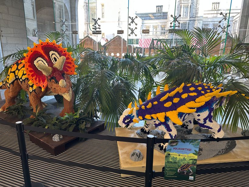 A large, colourful sculpture of a dinosaur stands on display, created from various LEGO blocks. The dinosaur, resembling a Triceratops, is detailed with vibrant patterns in red and orange and features a striking frill. Next to it, another dinosaur model, Polacanthus, is depicted with blue, yellow, and purple hues, surrounded by tropical plants. A sign is visible with information about Polacanthus.