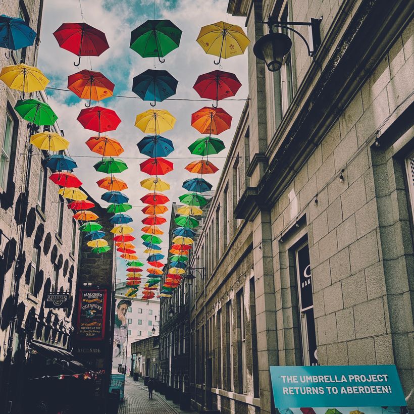 A vibrant display of colorful umbrellas hangs above a narrow street, creating a lively atmosphere. People walk beneath the umbrellas, which are suspended by wires. Stone buildings line the street, with one featuring a sign that reads, 'THE UMBRELLA PROJECT RETURNS TO ABERDEEN!' The sky is partly cloudy, adding to the cheerful ambiance.