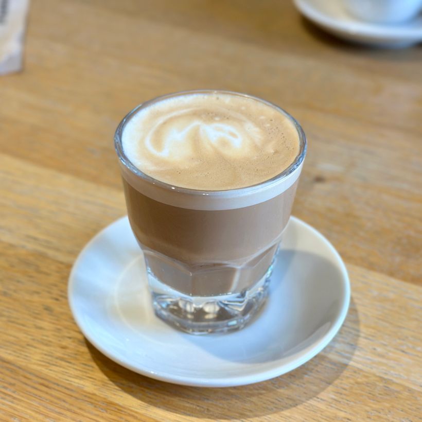 A glass filled with a layered coffee drink sits on a white saucer. The drink features a light brown top with a creamy froth, showcasing swirling patterns. It’s placed on a wooden table, with a blurred background hinting at a casual café setting.