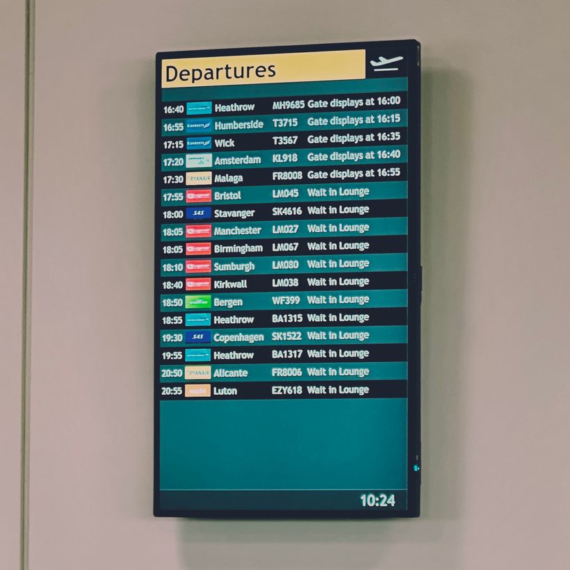 A departure board displays a list of flights along with their times, destinations, airlines, and boarding statuses. Flights to various locations, such as Heathrow and Malaga, are listed with scheduled times ranging from 16:40 to 20:55. The current time on the board shows 10:24.