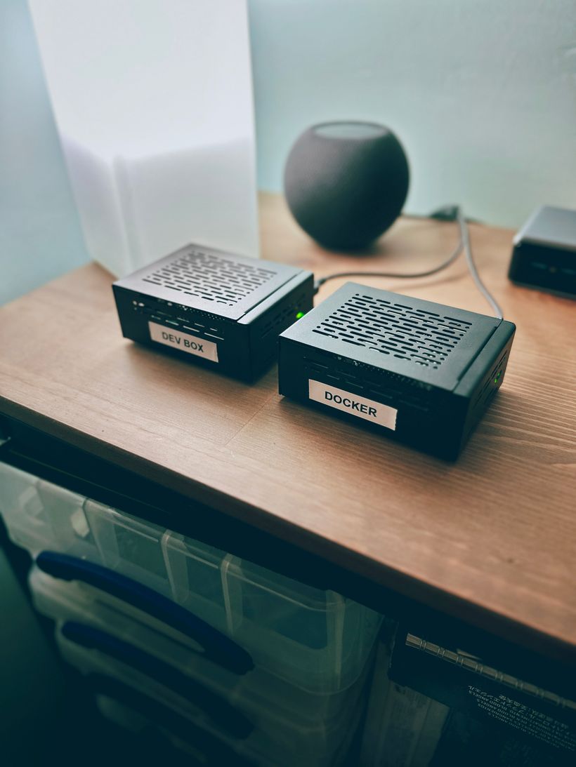 Two raspberry pi single-board computers, in small, black, metal chassis. One is labelled 'Dev Box', the other is labelled 'Docker'. There is a power cable connected to each, and green status lights indicate they are powered on. They sit on a wooden surface. There is a table lamp next to them, and an Apple Homepod speaker behind them.