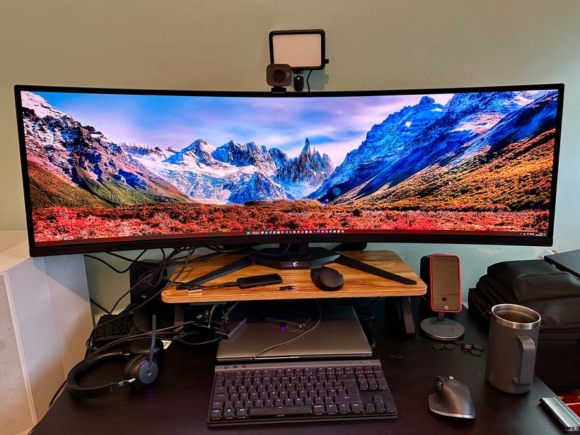 A very large, 49-inch curved computer monitor displays a vibrant landscape of mountains and a colourful foreground. Below the monitor, a wooden shelf holds a keyboard, mouse, and a headset. Nearby, there’s a silver mug and a smartphone on a stand, with a laptop and a backpack in the background.