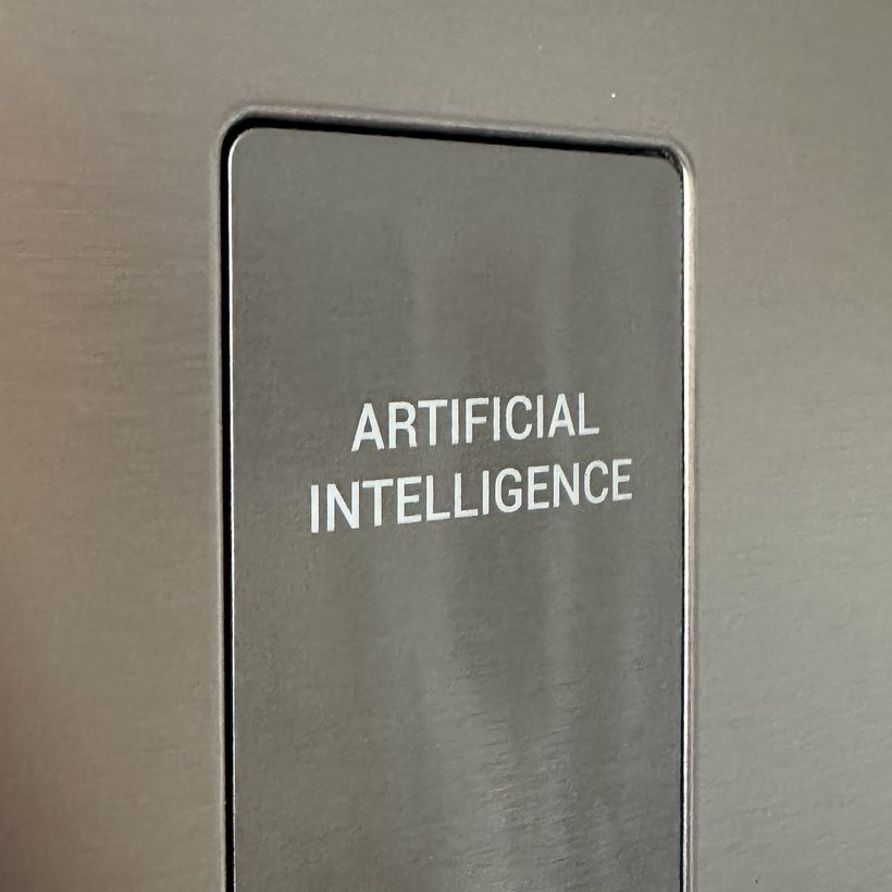 A rectangular metallic panel prominently features the text “ARTIFICIAL INTELLIGENCE” in bold, white letters. The panel appears sleek and modern, set against a darker metallic background.