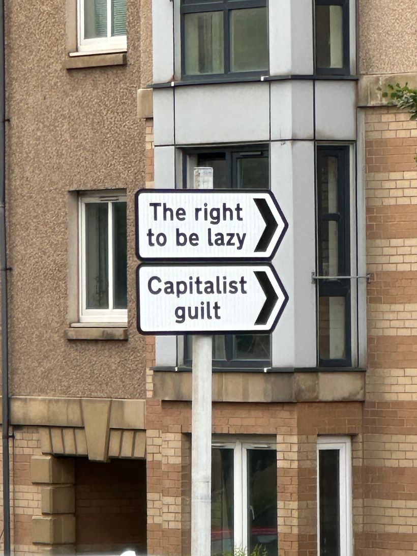 A street sign displays two directions with humorous phrases: 'The right to be lazy' pointing in to the right of the photo, and 'Capitalist guilt' pointing in the same direction. The sign is mounted on a pole near a multi-story building with a mix of textured walls and windows, reflecting an urban context.