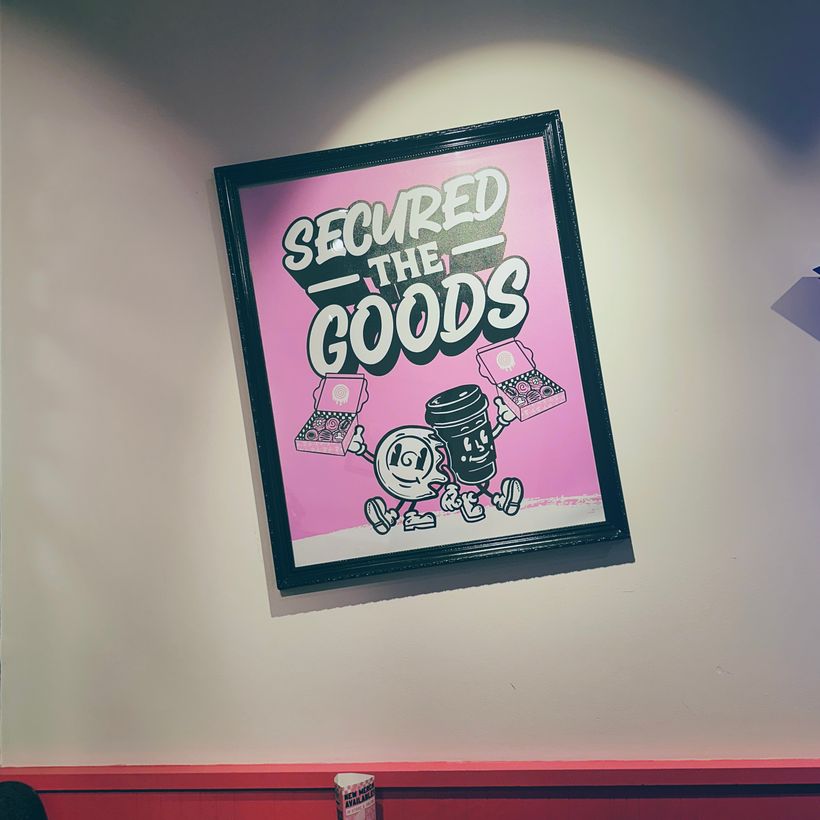 A framed poster displays a playful cartoon-style image featuring two characters: a round, white cinnamon roll and a black cup. They stand together, smiling, with pastry boxes on either side. Bold text above them reads, 'SECURED THE GOODS,' against a vibrant pink background, creating a cheerful dining atmosphere.