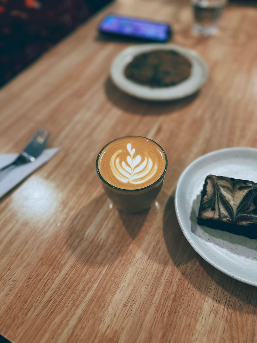 A small glass of cortado (a layered coffee drink made with espresso and milk) with a decorative leaf design sits on a wooden table. Nearby, a plate holds a brownie, and a round cookie is visible in the background, along with a smartphone and a partially visible glass of water, creating a cozy café atmosphere.